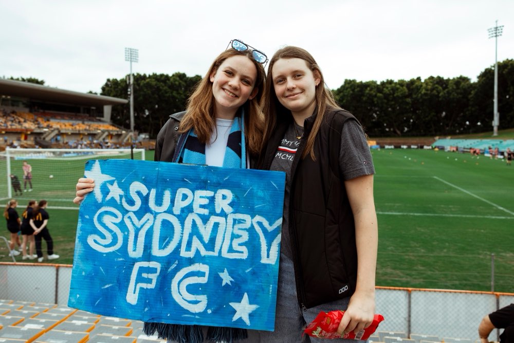 Two fans with a Super Sydney FC sign beside the pitch