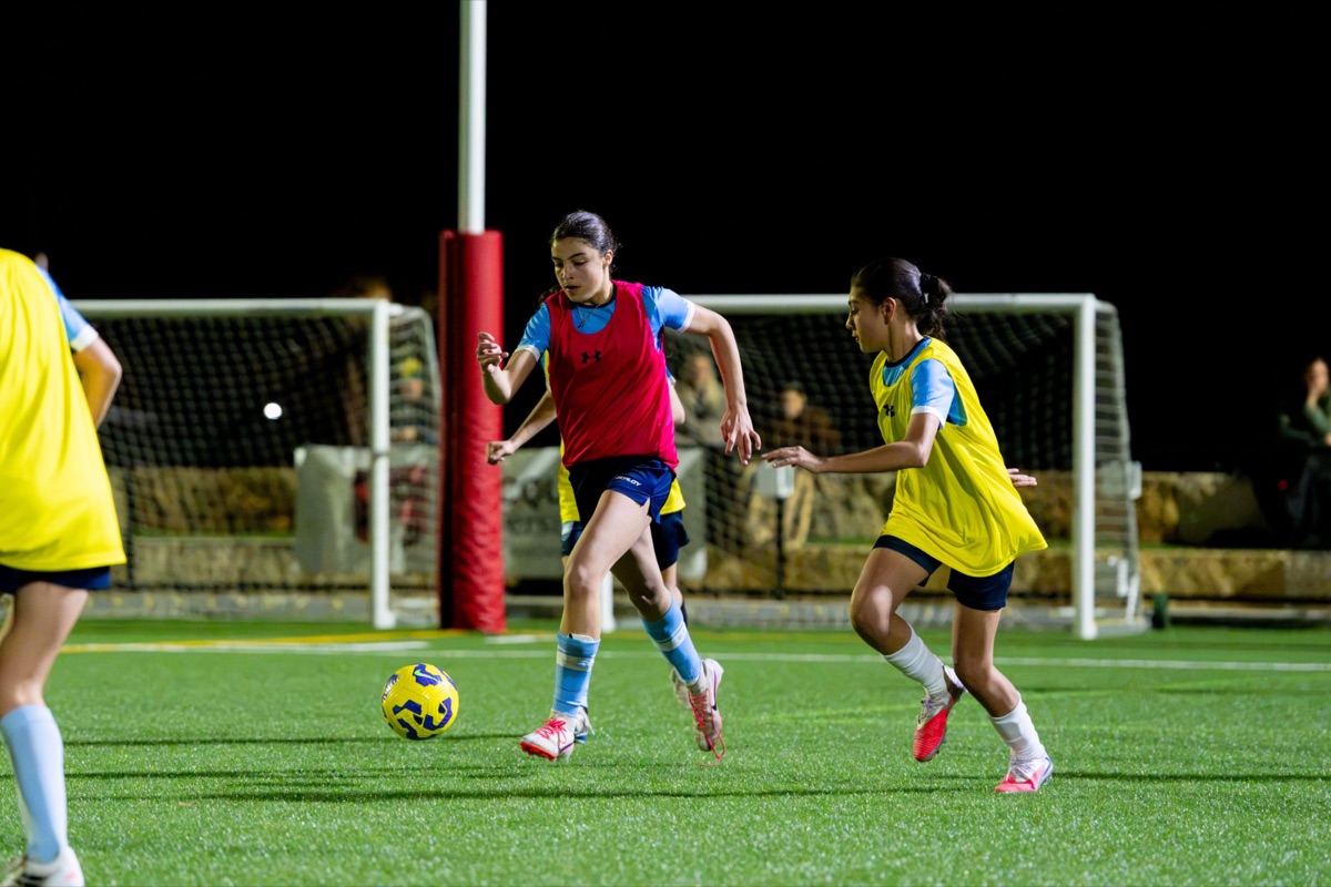 Two girls in training bibs chasing the ball under lights at a Sydney FC Female Training Program session